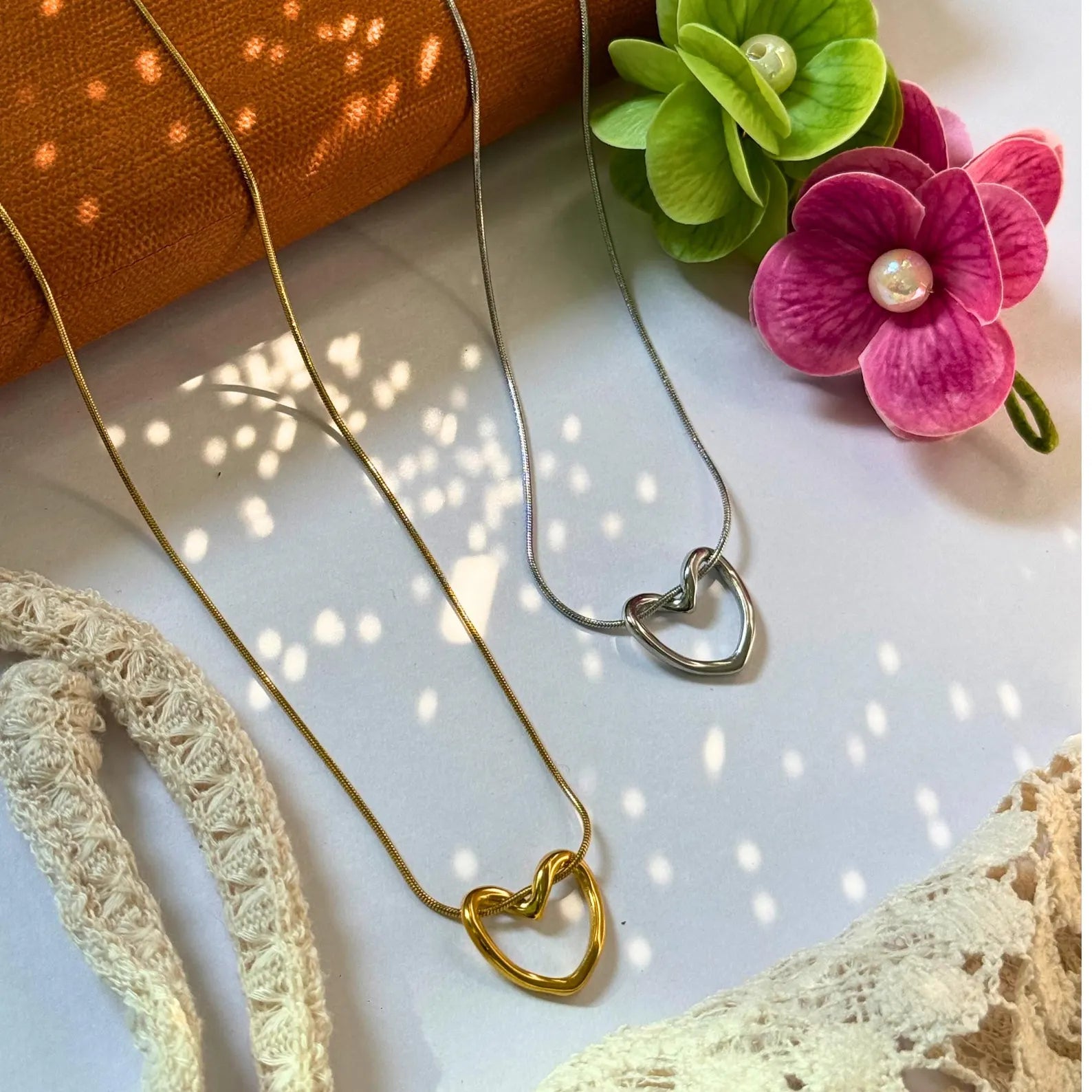 Two heart-shaped necklaces on a textured surface with flowers and lace.